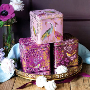 Three decorative metal tea or candy tins stacked on a round tray, featuring pink and purple floral and bird illustrations, styled with flowers and meringue on a wooden surface.