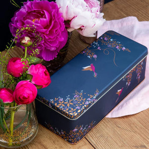 The Sara Miller Navy Hummingbird Rectangular Storage Tin next to pink flowers on a wooden surface