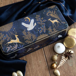 Lifestyle view of Magical Nightfall rectangular tin on wooden surface with treats and Christmas decorations.