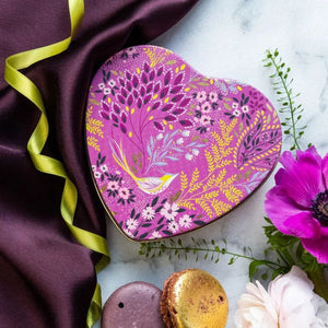 Sara Miller Mauve Songbird Small Heart Tin with a floral and bird pattern in pink, purple, yellow, and green, styled on a marble surface with a dark satin cloth, ribbon, flowers, and macarons.