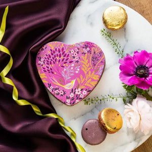 Sara Miller Mauve Songbird Small Heart Tin with a pink floral pattern sits on a white marble tray, styled with purple silk fabric, gold and pink flowers, and macarons.