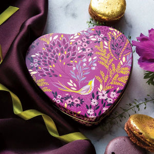 Sara Miller Mauve Songbird Small Heart Tin with a floral and bird pattern in purple, pink, yellow, and white, styled on a marble surface with purple fabric, macarons, and flowers.