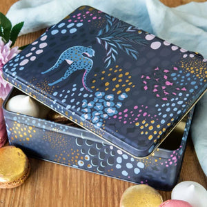 Sara Miller Midnight Leopard Biscuit Tin decorated with a blue leopard and colorful dotted botanical patterns, shown open on a wooden surface with macarons and soft fabric around it.