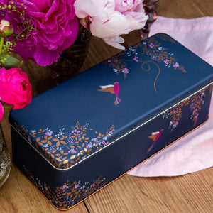 The Sara Miller Navy Hummingbird Rectangular Storage Tin on a wooden surface with flowers.