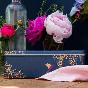 The Sara Miller Navy Hummingbird Rectangular Storage Tin on a wooden surface with flowers in the background