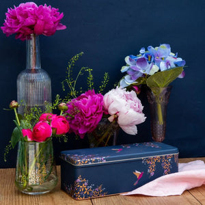 Sara Miller Navy Hummingbird Rectangular Storage Tin surrounded by flowers on a wooden surface.