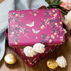 Sara Miller Orchard Butterfly Biscuit Tin shown open with white meringues inside on a wooden surface, with macarons and flowers nearby.
