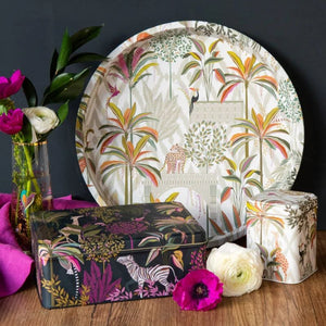 A styled product display featuring a large round tray and two decorative boxes with vibrant tropical and safari-themed prints by Sara Miller, arranged on a wooden surface with flowers against a dark background.