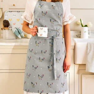 A woman wearing the Sophie Allport Chicken Apron with a colourful chicken and white dot pattern, holding a white mug with a chicken illustration, in a bright kitchen setting.