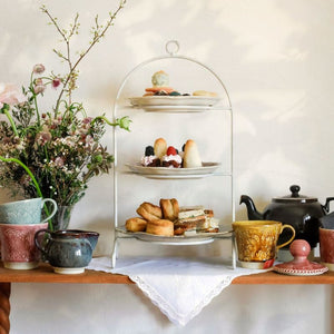 Three-tiered cake stand with pastries, teacups and creamer from the Sthal Arabesque range, and a vase of flowers on a wooden surface.