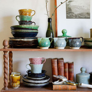 Wooden shelf with Sthal Arabesque ceramic plates, cups, bowls, and books against a neutral wall.