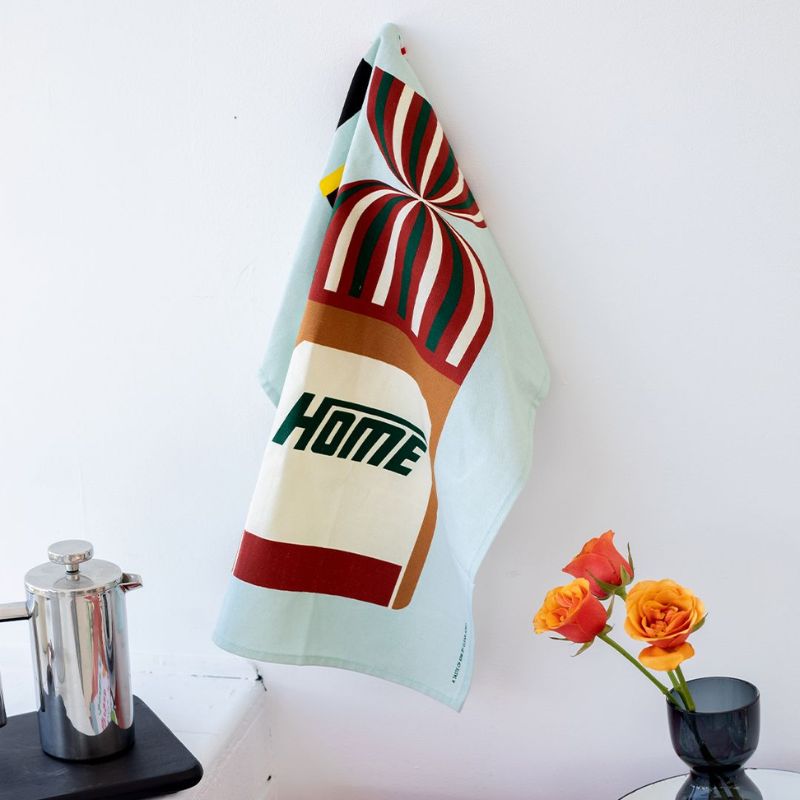 100%NZ Taste of Kiwi Tea Towel hanging from a hook in a kitchen - Vogels bread icon showing.