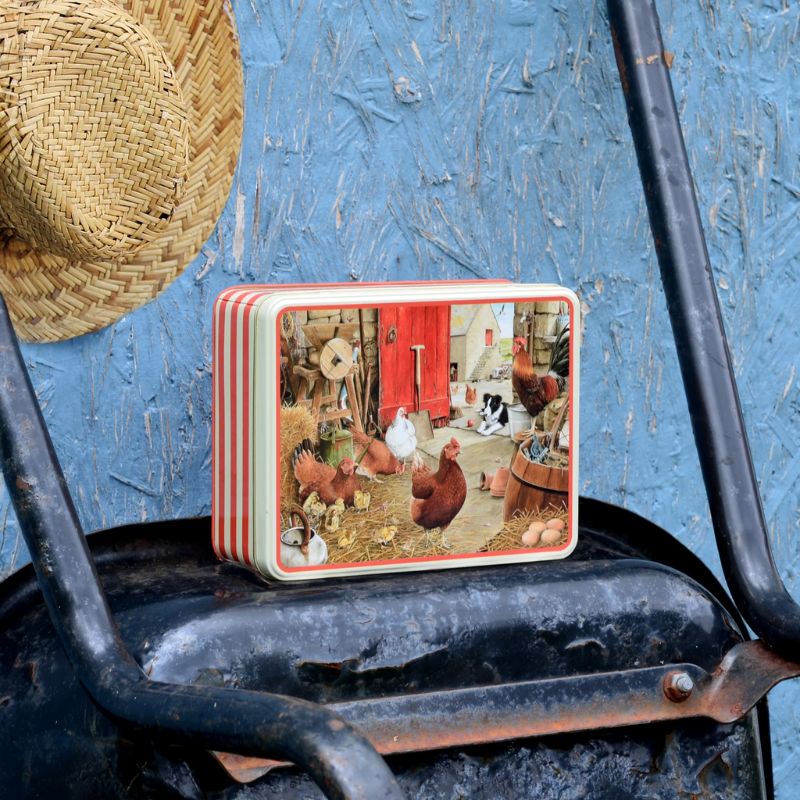 The Tracy Hall Farmyard Hens Biscuit Tin placed on a wheelbarrow with a straw hat behind
