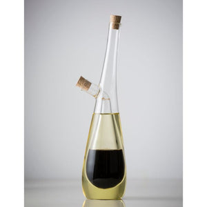 A clear glass oil dispenser bottle with a cork stopper and a side branch, partially filled with oil and vinegar, shown against a light gray studio background.