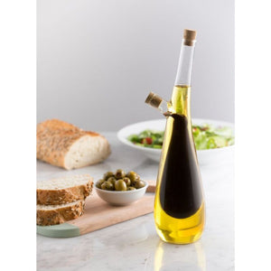 The Typhoon Tear Drop Glass Duo Drizzler filled with oil and vinegar set on a light marble surface with bread, olives, and a salad bowl in the background.