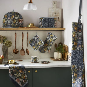A kitchen scene styled with a dark navy floral apron hanging on the right, matching oven mitts and a towel from the Ulster Weavers Finch & Flower collection. Styled on a wooden shelf, along with copper utensils, a mug, and decorative items on a white countertop.