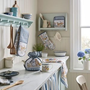 A kitchen scene featuring a coordinated set of blue and white gingham and floral-patterned kitchen towels, oven mitts, and teapot covers from the Ulster Weavers Pantry Cats collection, styled on a marble countertop and hanging hooks.