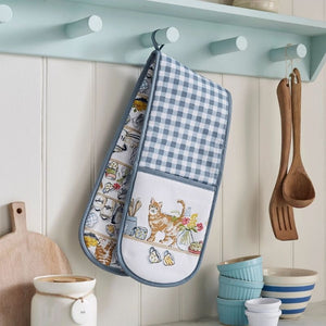 A blue and white gingham oven mitt with a printed cat themed pocket hangs on a kitchen peg rack. Wooden utensils hang on the right, and bowls and a cutting board are visible on the counter below.