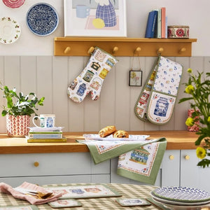 A styled kitchen scene with two patterned oven mitts hanging on pegs and matching dishcloths draped over a wooden counter from the Ulster Weavers Tea Tins collection.