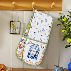 Ulster Weavers Tea Tins Double Oven Glove hanging from wooden wall hooks. The glove is white with blue floral prints and a large blue-and-white tea jar illustration on the front pocket, with green trim.