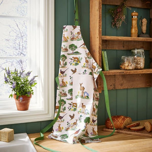 Ulster Weavers Thistlewood Cotton Apron hanging on a green wall next to a window with a plant, wooden shelves with jars, and bread.