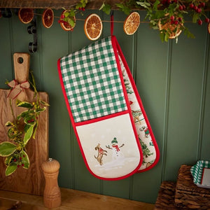 Ulster Weavers Winter Walkies Double Oven Glove hanging on a dark green paneled wall, decorated with a green and white gingham pattern, red trim, and a white lower section featuring a snowman and dog illustration. Dried orange slices, greenery, and holiday garlands hang above.