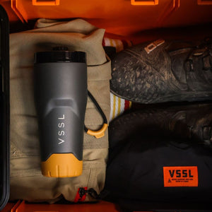 VSSL Rift Tumbler - Ash with a dark gray body and yellow-orange accents, resting in a packed orange storage compartment alongside hiking shoes.