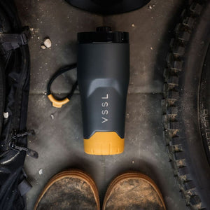 VSSL Rift Tumbler in matte dark gray with a mustard yellow base, lying on a dirt surface next to a black backpack and tire, with the wearer's boots visible at the bottom.