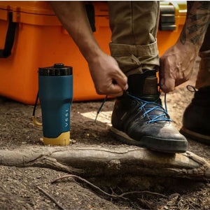 VSSL Rift Tumbler in Pacific Blue next to a person tying the laces of a black hiking boot. The person is wearing khaki pants, and an orange cooler is visible in the background.