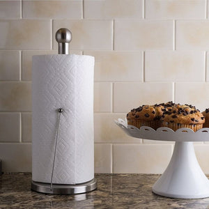 A roll of white paper towels on the Westmark Kitchen Roll Holder, set on a kitchen counter with a tiled backsplash and chocolate chip muffins on a white cake stand.